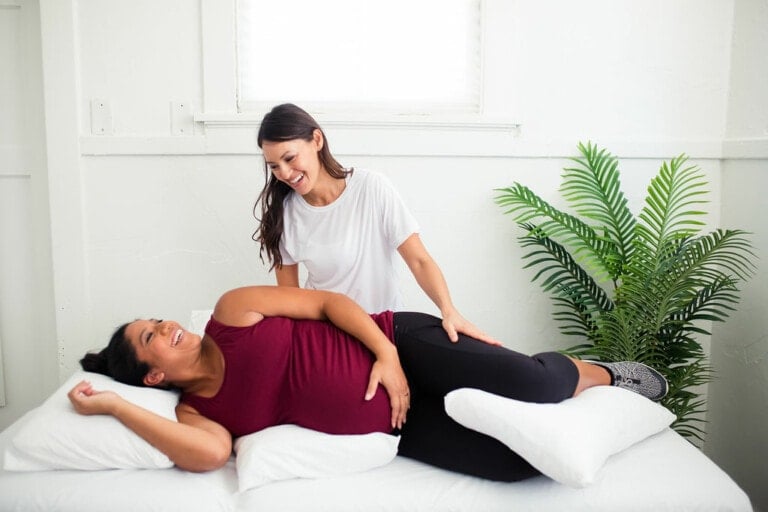 A woman in a purple tank top lies on her side on a bed, supported by pillows. Another woman in a white shirt smiles while gently pressing on her back, suggesting ways to heal your pelvic floor. There is a green potted plant in the background.