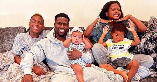A family of five sits on a couch, smiling for the picture. Two adults and three children are present, with the celebrity dad holding two of the children while the other child stands to the side, posing. They are casually dressed and the background is a plain wall.