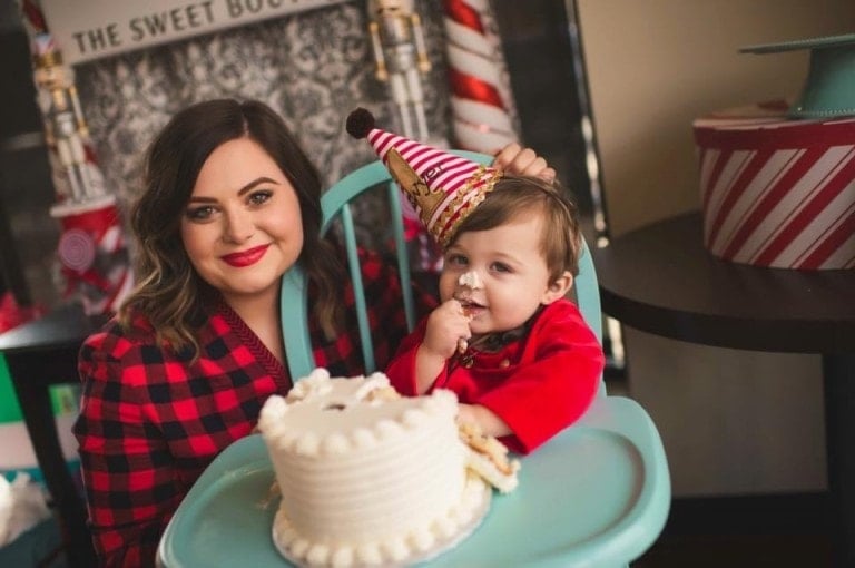 A woman and a child are seated together in front of a white cake at a cheerful birthday soirée. The child, wearing a party hat, has a piece of cake in his hand and icing on his face. The woman, smiling and dressed in a red plaid shirt, enjoys the moment amidst the festive decorations in the background.