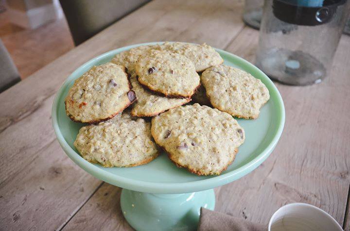 A green pedestal cake stand holds a batch of oatmeal lactation cookies with chocolate chips, placed on a wooden table. The background features a blurred home interior with a glass jar and a cup partially visible.