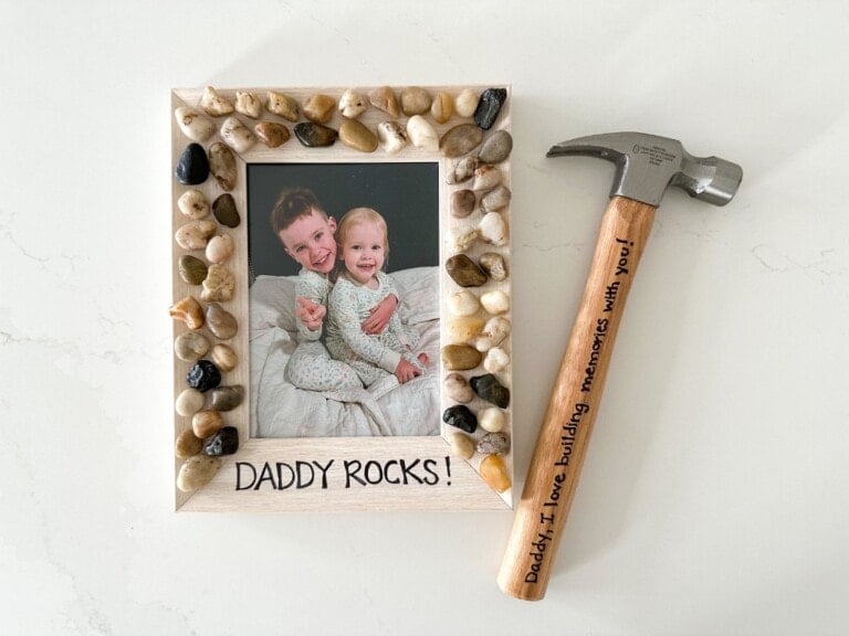 A framed photo featuring two children, decorated with rocks and the text DADDY ROCKS! Additionally, a hammer with an inscription on the handle that reads, Daddy, I love building memories with you! is placed beside the frame. Perfect for Father's Day crafts!