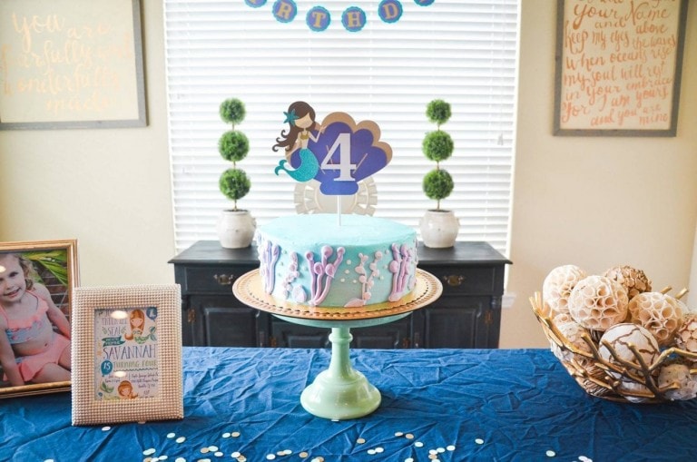A round, decorated cake with a "4" topper and colorful ocean-themed decorations sits on a green cake stand on a blue tablecloth. The table holds a framed photo of a child, a mermaid birthday sign, and decorative objects. Banners and framed quotes are on the wall.