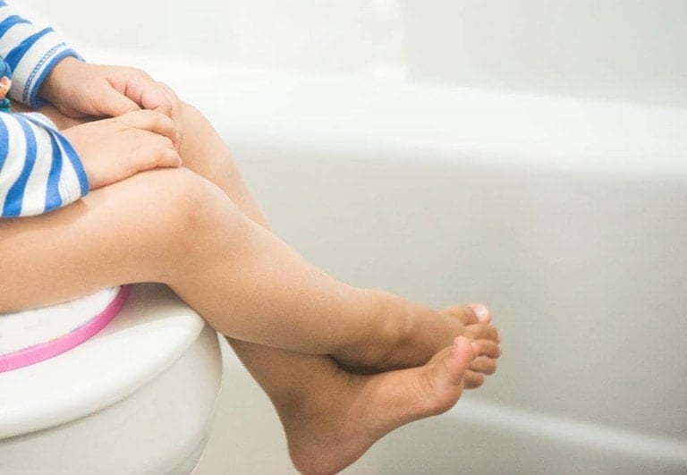 A child is sitting on a toilet seat with legs crossed, wearing a long-sleeve shirt with blue and white stripes, showcasing their potty training progress. The background reveals part of a white bathtub and bathroom wall.