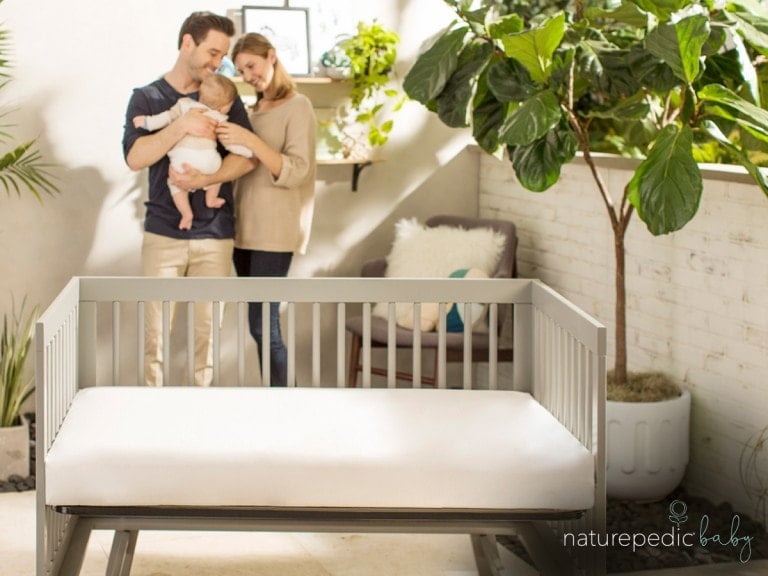 A couple stands in a well-lit room holding a baby. In the foreground, there is an empty gray crib with a white mattress—choosing the right crib mattress is an important decision for parents. Plants are visible in the background, and a logo in the bottom right corner reads "naturepedic baby.