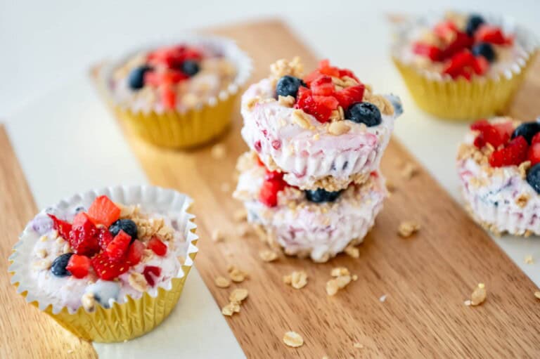 A wooden surface holds several yogurt cups in yellow paper liners, filled with a creamy mixture and topped with sliced strawberries, blueberries, and granola. Two yogurt cups are stacked in the center. The scene is bright and colorful—perfect inspiration for your next yogurt snacks adventure.