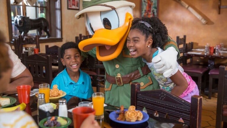 A person dressed as a cartoon duck in a safari outfit poses with two smiling children at a dining table. The children embrace the character while seated in a restaurant with plates of food and drinks on the table. The background shows a warm, casual dining setting, perfect for those using their Disney dining plan.