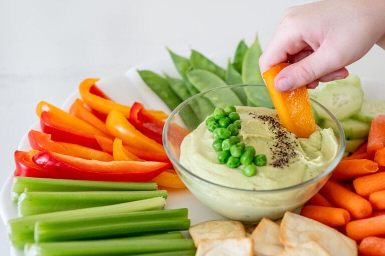 A hand dips a sliced orange bell pepper into a bowl of green pea hummus. The bowl sits on a platter surrounded by sliced red, yellow, and orange bell peppers, celery sticks, snow peas, cucumber slices, baby carrots, and pita chips—perfect for pairing with Stonyfield yogurt pouches or using in yogurt recipes.