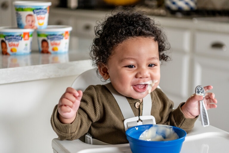 A smiling baby with curly hair sits in a high chair, wearing a brown long-sleeve shirt. The baby has Stonyfield yogurt on their face and holds a spoon in one hand. There is a blue bowl with yogurt on the high chair tray. In the background, containers of baby food are on the counter.