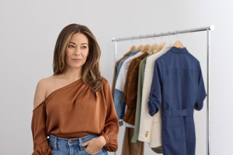 Sarah Villarreal stands near a clothing rack filled with various garments on hangers, wearing a brown off-shoulder blouse and blue jeans in a bright, minimal room.