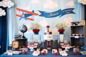 A birthday party setup with an aviation theme, featuring a backdrop with a red airplane and a banner reading "Joshua is turning One!" The table is decorated with cakes, flowers, a globe, and various treats labeled with food names. It's one of the most delightful first birthday themes around!