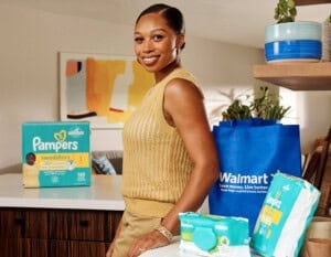 Allyson Felix stands in a kitchen, smiling at the camera. In front of her, on the counter, are packages of Pampers diapers, baby wipes, and a Walmart reusable shopping bag. Shelves with plants and bowls are in the background.