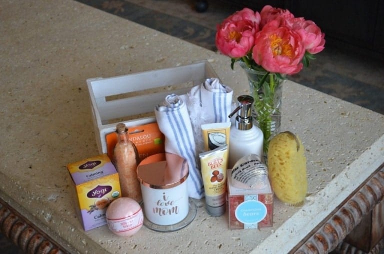 A white crate holds rolled white towels and sits on a countertop with various items around it, including tea boxes, lotion, a candle, bath bombs, a sea sponge, and a bouquet of pink flowers—perfect for creating a DIY Mother's Day spa in a box.