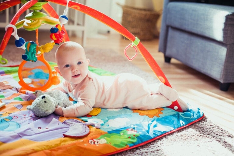 A baby in a white onesie lies on their stomach on a colorful play mat with some of the best developmental toys for babies attached overhead. The baby looks up with a smile, holding a gray stuffed animal. The room is well-lit with a gray piece of furniture visible in the background.