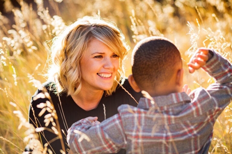 A woman with blonde, shoulder-length hair smiles at a child in a plaid shirt. They are outdoors surrounded by tall, golden grass. The woman is crouching, and the child is facing away from the camera, lifting his arms happily. The scene is brightly lit by the sun, capturing their ever after moment.