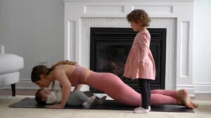 A woman is doing a push-up while looking at a baby lying beneath her. A young girl in a pink dress stands nearby. They are in a living room with a fireplace in the background.