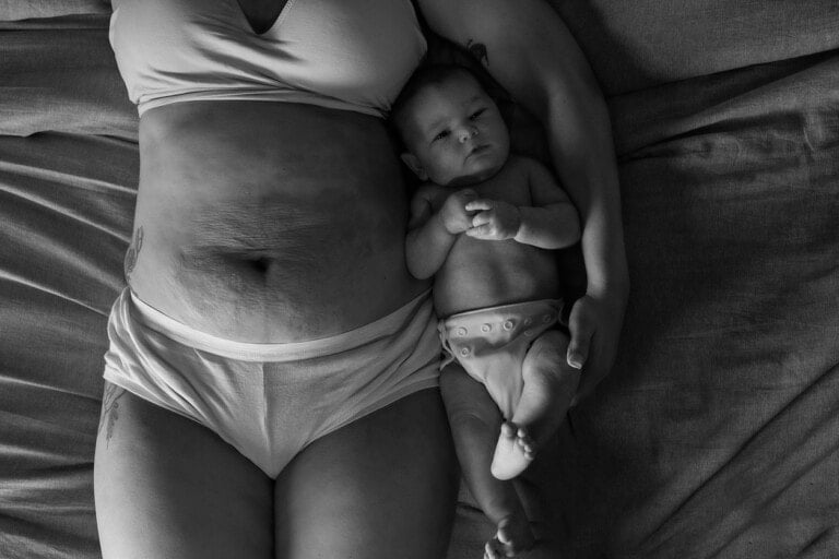 A black and white photo captures a tender moment of preparing for postpartum life: an adult lies on a bed with a baby. The adult, in light-colored underwear and bearing visible stretch marks, cradles the diaper-clad infant close with an arm.