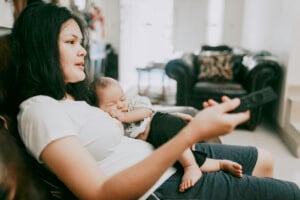 A woman sits on a couch, holding a remote control in her right hand and a sleeping baby on her lap. She wears a white t-shirt and dark shorts. The living room in the background has another couch and various household items, as she considers shows to binge-watch on maternity leave.