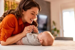 A woman with long brown hair, wearing an orange blouse, smiles and holds the hands of a 2-month-old baby lying on a white surface. The baby is dressed in a gray outfit. They are in a bright, modern indoor setting.