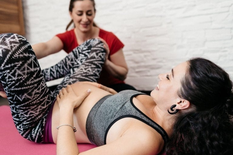 A pregnant woman in exercise clothing is lying on a yoga mat with one hand on her belly. Another person, wearing a red shirt, is assisting her with a leg stretch. They are indoors, with a white textured wall in the background, likely preparing for their upcoming doula podcast.