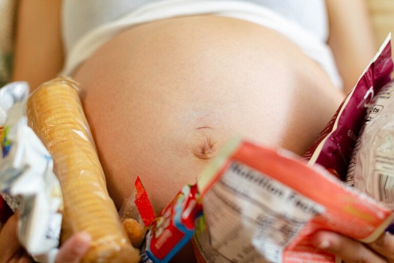 A close-up photograph captures a pregnant belly surrounded by various snack food items, including a pack of biscuits, a bag of chips, and other packaged snacks—perfect evidence of pregnancy cravings. The person is wearing a white top.
