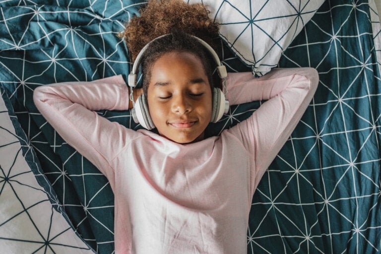 A young girl with curly hair is lying on a bed with a geometric patterned comforter, wearing headphones and a pink long-sleeved shirt. Her eyes are closed, and she appears to be relaxed, enjoying one of her favorite quiet time activities.