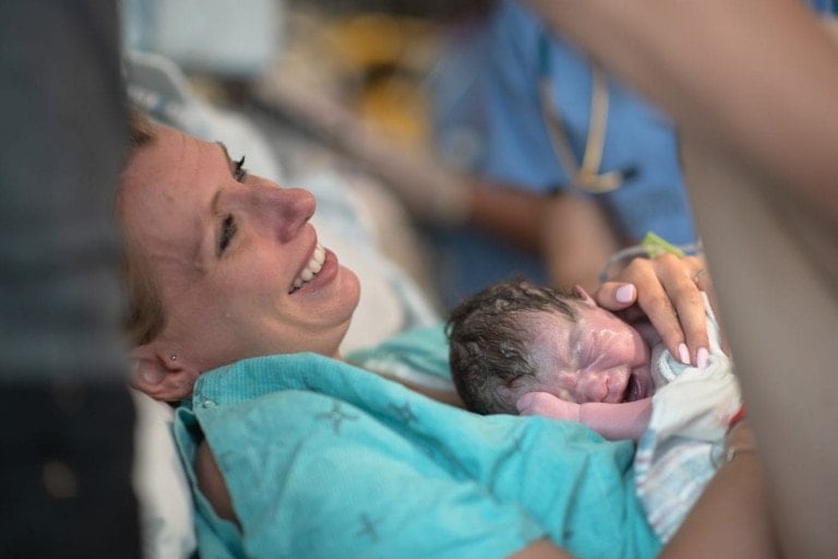 A woman in a hospital gown smiles while holding a newborn baby. The woman is lying in a hospital bed, and a person in blue medical scrubs is partially visible in the background, suggesting a hospital setting. Despite the challenges of perineal tearing, her joy is palpable.