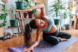 A pregnant woman practices yoga on a mat in a bright room. She is seated in a side stretch position, smiling. Several potted plants are in the background, and the floor is wooden.