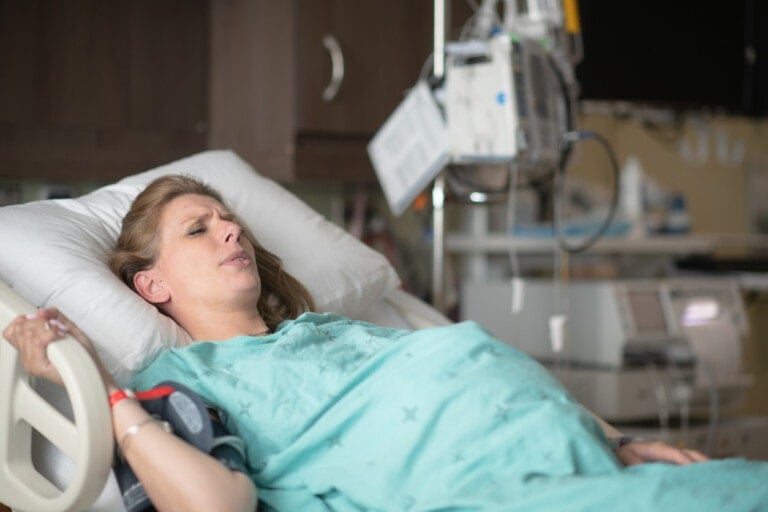 A woman in a hospital bed, wearing a teal hospital gown, appears to be in labor. She is lying on her back, holding her arm, with medical equipment and monitors visible in the background, possibly preparing for a Foley bulb induction procedure.