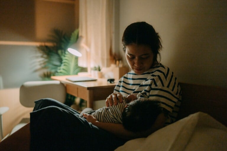 A person in a striped shirt cradles a baby in their arms while sitting on a bed in a dimly lit room. A desk lamp illuminates the background, casting light on a plant near the desk. The scene appears calm and intimate, quietly reflecting concerns of low milk supply.