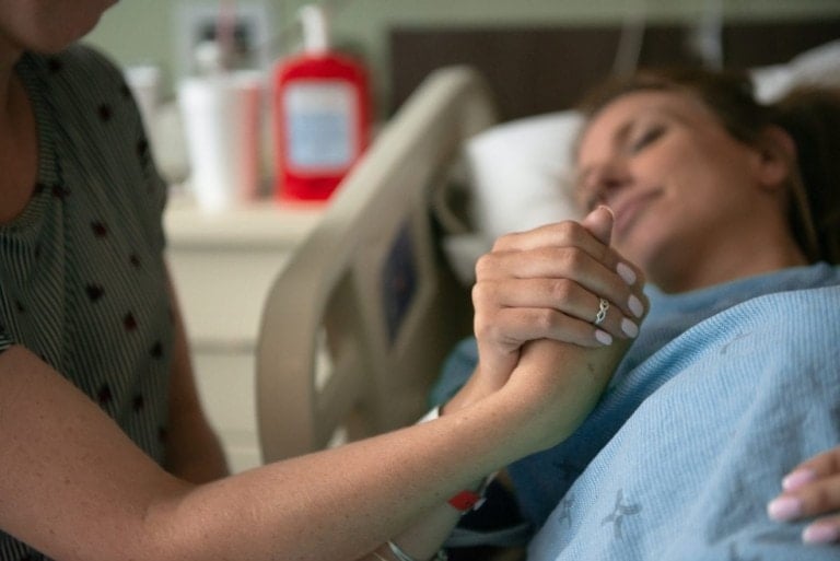 A person lying in a hospital bed holding hands with another, who sits beside them. The patient, in a blue hospital gown, is surrounded by various medical items. The tender moment echoes the Love Languages of Labor and Delivery, showcasing the bond and support during challenging times.