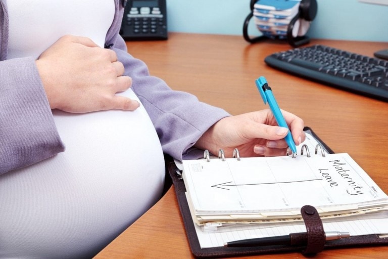 A pregnant person wears a purple cardigan and sits at a desk, holding their belly with one hand and writing "Maternity Leave Plan" in a planner with the other. A computer keyboard and office supplies are visible in the background.