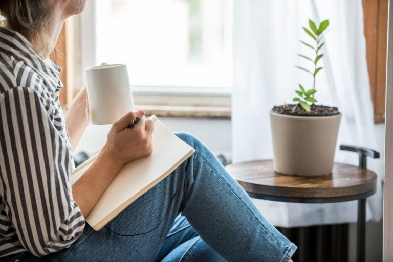 A person in a striped shirt and jeans is sitting by a window, holding a cup and writing in a notebook, fostering a positive mindset. A small potted plant is on a round wooden table next to them. Light filters through a white curtain, creating a serene atmosphere.