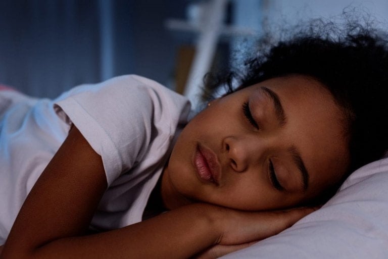 A young child with curly hair is sleeping peacefully on their side, aided by melatonin safe for kids. The child rests their head on a pillow, eyes closed, and wears a white t-shirt. The background is softly lit, creating a calm and serene atmosphere.