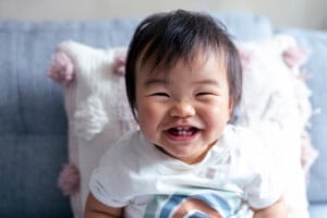 A smiling baby with dark hair and a white shirt sits on a light blue couch, celebrating their 6-month milestones. The baby is in focus while the background is slightly blurred. A white pillow with decorative tassels is behind the baby.