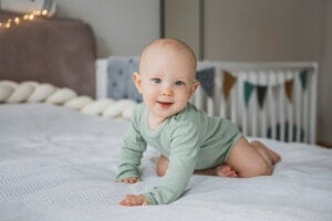 A baby wearing a light green onesie is crawling on a bed with a white blanket. The baby, perhaps named one of those popular boy names that start with W, has a slight smile and looks towards the camera. In the background, there is a crib decorated with a string of triangular flags.