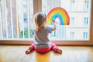 A young child with blonde hair, wearing a blue polka dot shirt and pink pants, sits on a wooden floor by a window. The child holds up a colorful drawing of a rainbow to the window, with buildings in the background, perhaps imagining unique rainbow baby names for each hue.