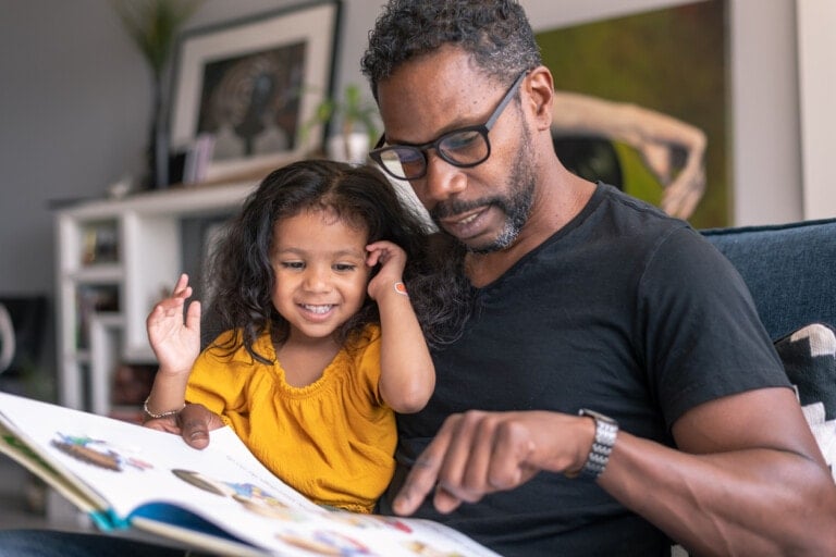 A man wearing glasses and a black t-shirt reads a book to a young girl in a yellow dress. Seated together on a couch in the living room, they cherish the little moments as the girl smiles and looks at the book while the man points to something on the page.