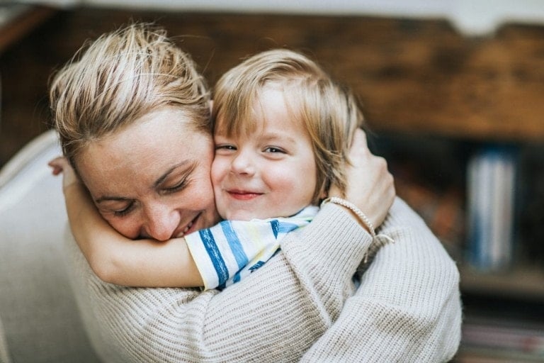 A woman with blonde hair hugs a smiling child with light brown hair, radiating kindness and compassion. The woman has her eyes closed and is wearing a light-colored sweater. The child, dressed in a striped shirt, looks at the camera. They are indoors with a blurred background, making the moment feel intimate and warm.