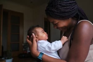 A woman smiles warmly while holding a baby in her arms. The baby looks up at her, and they make eye contact, sharing a tender moment of baby bonding indoors.