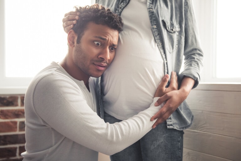 A man with a surprised expression kneels and puts his hands and ear on a pregnant woman's belly, eagerly listening for baby news. The woman, wearing a white shirt and denim jacket, stands beside him. They are indoors with light streaming through the window behind them.