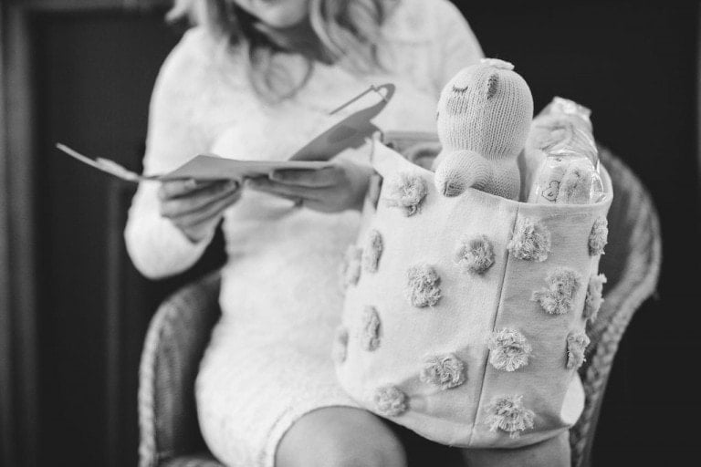 A person in a white lace dress reads a card while sitting on a chair. In their lap is a textured fabric basket containing a knitted bear toy and two baby bottles, perhaps gifted during the joyous occasion—a perfect snapshot for baby shower hashtags. The scene is in black and white.