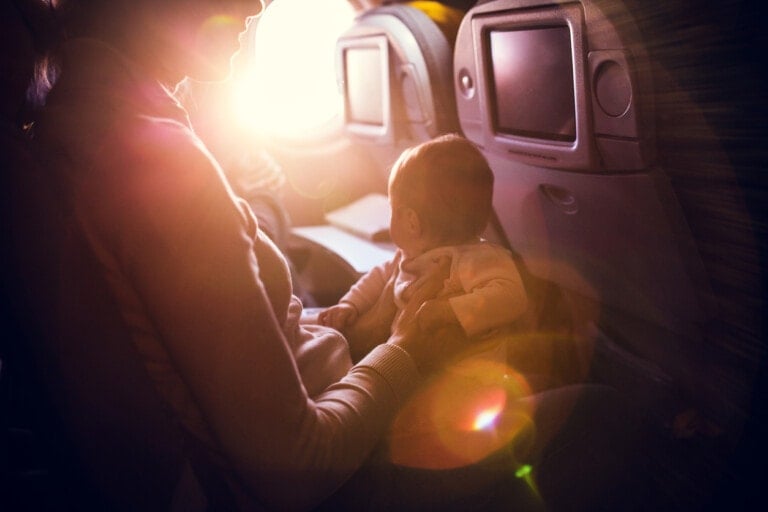 A person is flying with a baby on their lap while sitting on an airplane. Sunlight is streaming through the window, creating a warm glow inside the cabin. Two backseat entertainment screens are visible on the seats in front of them.