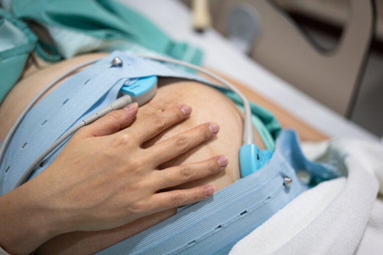 A close-up shows a pregnant person lying down with their hand resting on their belly. Medical monitoring equipment, including a fetal heart rate monitor, is strapped around their abdomen. The individual is wearing a hospital gown as they prepare for labor induction, and a bed is partially visible.