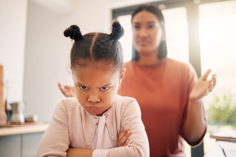 A young girl with arms crossed and an obstinate expression stands in the foreground. In the background, an adult woman appears exasperated, her arms raised slightly. They are indoors with a bright window behind them.
