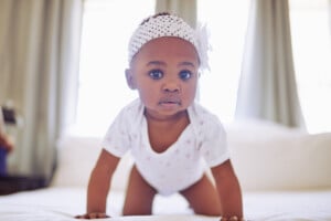 A baby girl named Olivia, wearing a white onesie and a white knitted headband with a flower, is crawling on a bed. The background consists of neutral-colored curtains and a bright window behind the child.