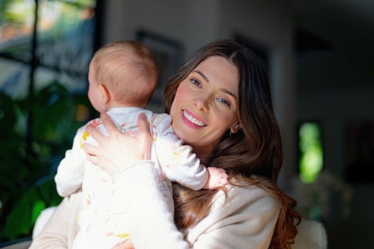 Ashley Greene, with her long brown hair, is smiling at the camera while holding a baby who is turned away. Indoors, plants and blurred background scenery add a serene touch. Ashley is wearing a light-colored top and the baby sports light-patterned clothes.