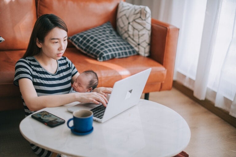 A woman is sitting at a white table, working on a laptop while holding a baby. A smartphone and a cup are on the table. She is seated in front of a brown sofa with cushions. Natural light is coming through a window with white curtains in the background, creating an inviting space free of unsafe products.