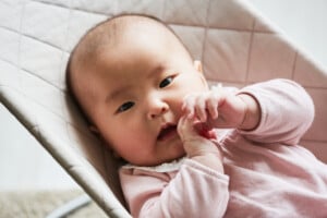 A baby girl in a light pink outfit lies on a quilted surface. She has a calm expression and appears to be touching her mouth with her hands. The background is softly blurred, emphasizing the baby as the focal point of the image, perfect for parents considering V girl names like Violet or Vivian.