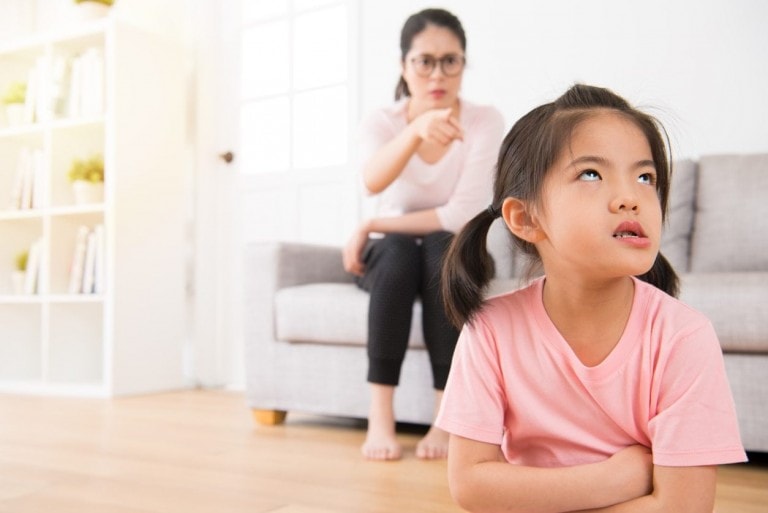 A young girl with pigtails and a pink shirt sits on the floor looking up with an annoyed expression. An adult with glasses sits on a couch behind her, pointing and appearing to scold the entitled child. The background shows a bright, neatly arranged living room.
