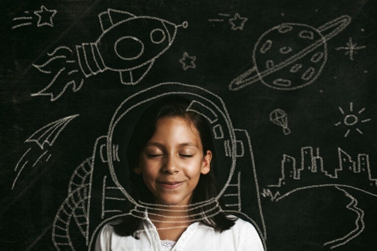 A young girl with closed eyes smiles in front of a chalkboard. Chalk drawings of a spaceship, planet, stars, and city skyline are behind her, with a chalk-drawn spacesuit outlined around her, giving the illusion she is wearing it. This scene could easily be featured on an episode of the Empower Our Girls podcast.